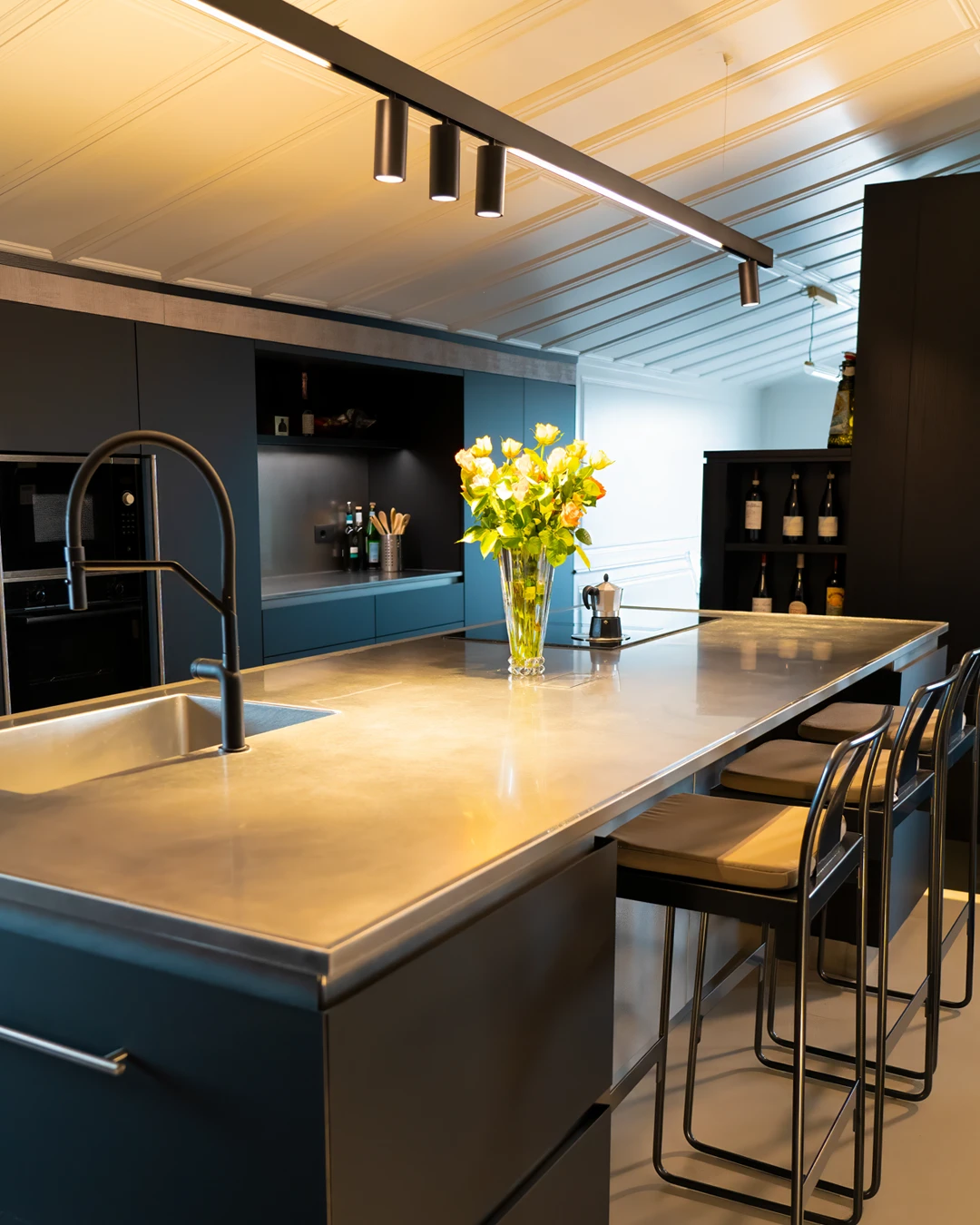 Cucina dal design moderno e industriale con ampia isola centrale dotata di top in acciaio inox, rubinetteria nera opaca, sgabelli da bar e mobili scuri. Sul piano di lavoro spicca un vaso di rose gialle illuminato da faretti a binario.